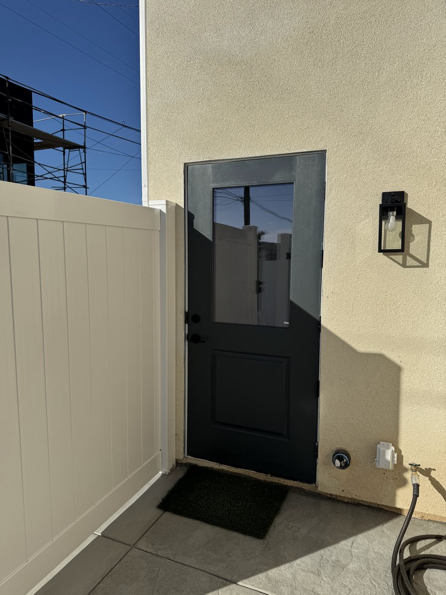 Black exterior door with frosted glass panel beside a white vinyl fence gate on a stucco wall with a modern wall sconce.