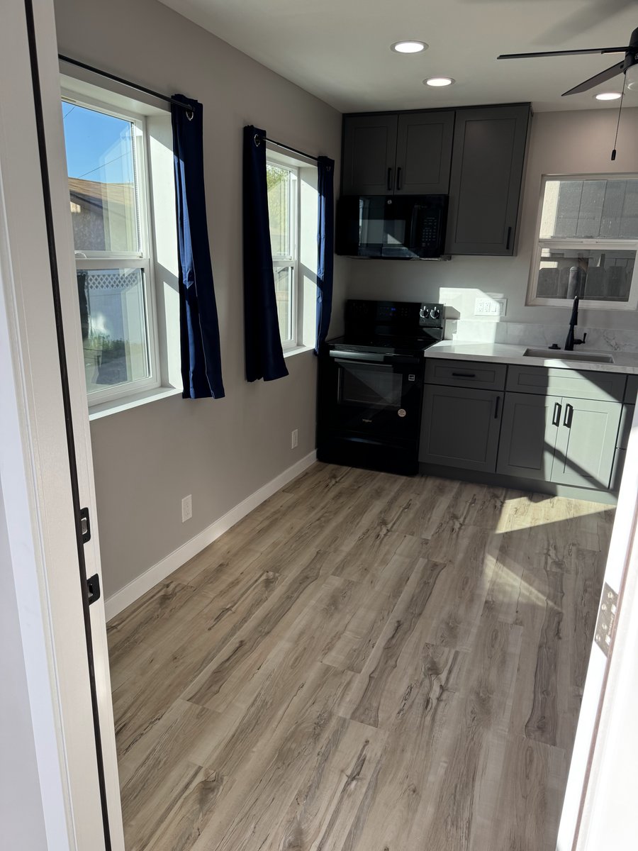 Interior view looking into a modern ADU kitchen with gray shaker cabinets, black appliances, white countertops, recessed lighting, wood-look vinyl flooring, and two windows with navy curtains.