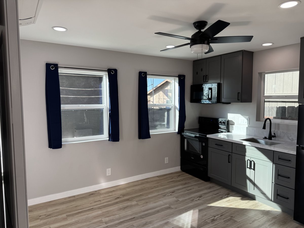 ADU kitchen and living area with two windows and navy curtains, gray cabinets, black appliances, white countertops, recessed lighting, a black ceiling fan, and wood-look flooring