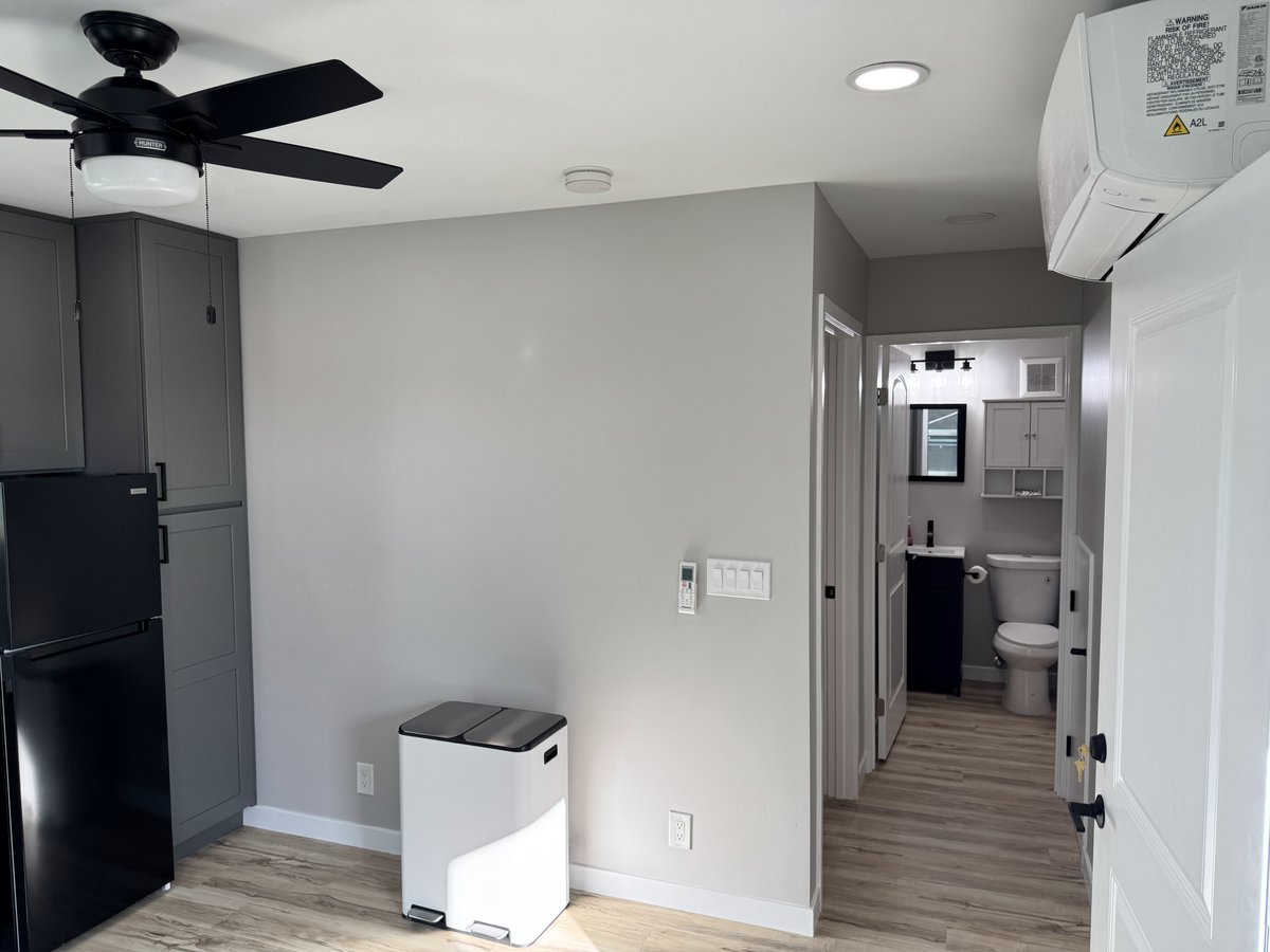 ADU interior view showing gray walls, LVP flooring, a ceiling fan, black refrigerator, wall-mounted mini split unit, and a hallway leading to the bathroom.