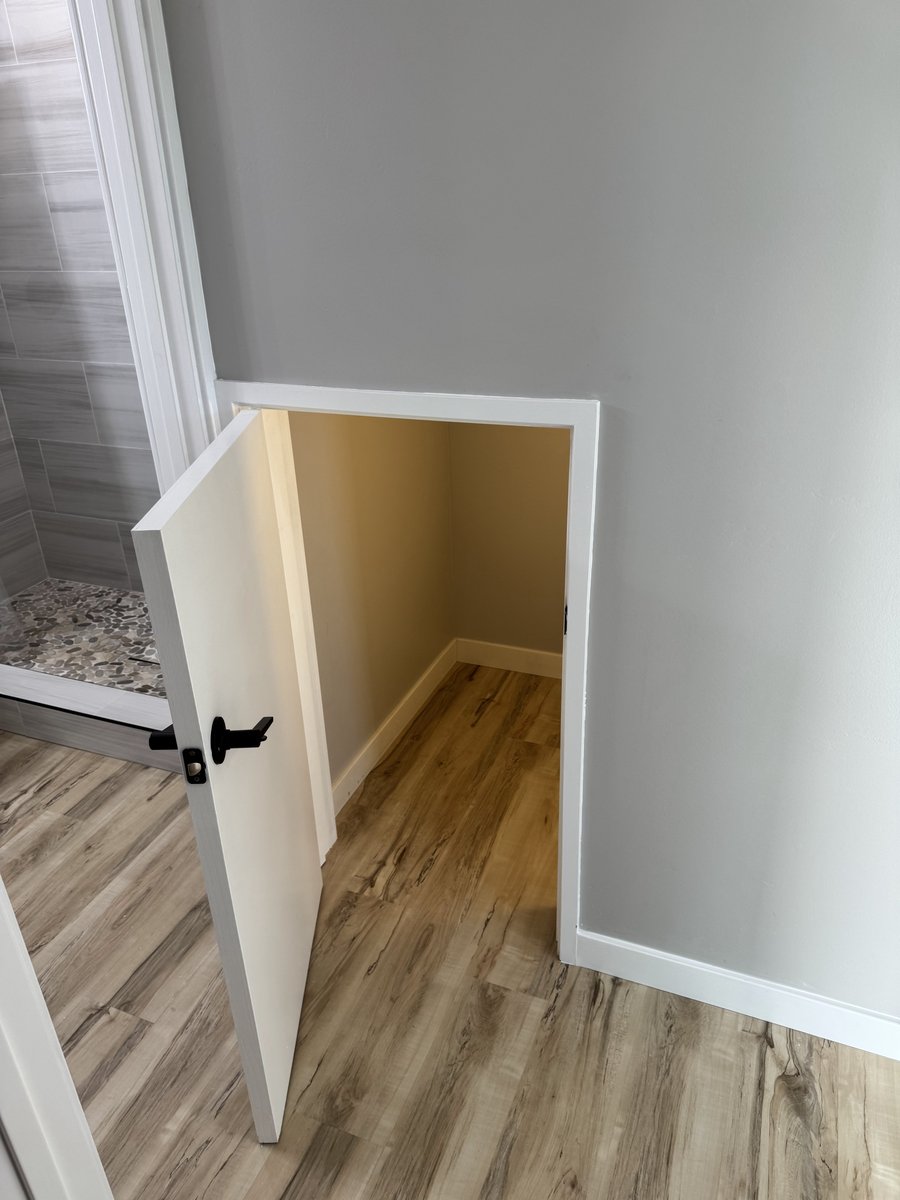 Small under-stair storage closet with a white door open, showing wood-look flooring and beige interior walls next to the bathroom entry.