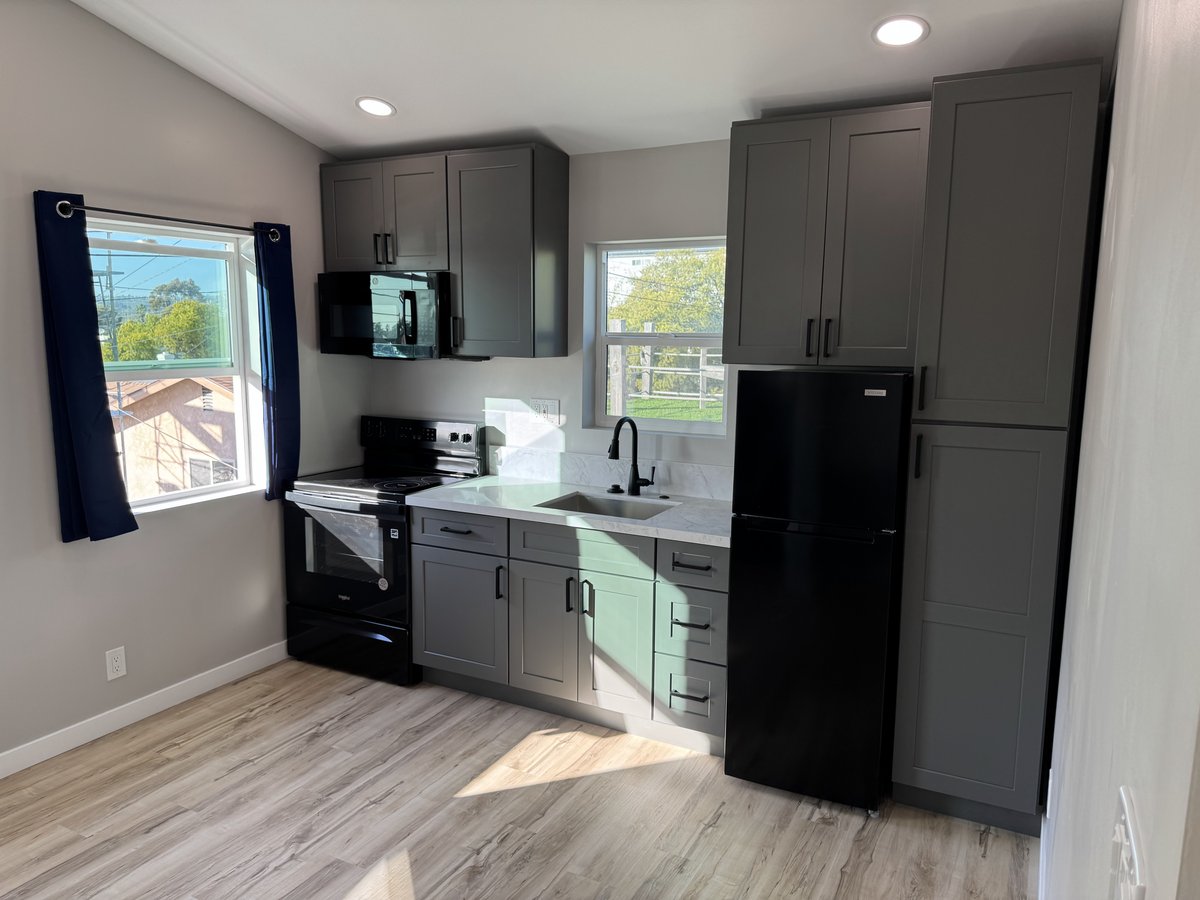 Small ADU kitchen with gray shaker cabinets, black refrigerator, black stove and microwave, white countertops, stainless sink under a centered window, and wood-look flooring with sunlight coming in.