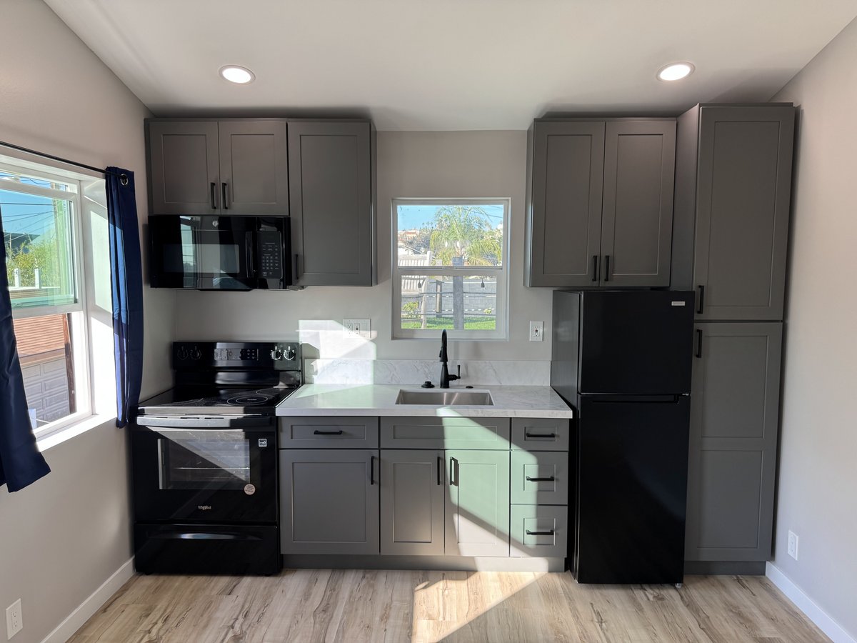 Compact ADU kitchen with gray shaker cabinets, white quartz countertops, black stove and microwave, black refrigerator, and a single-basin sink under a centered window.