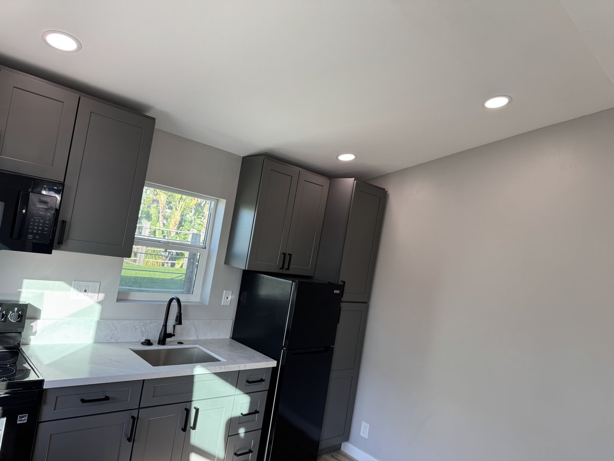 Modern ADU kitchen corner with dark gray shaker cabinets, white quartz counters, black sink and faucet, and a black refrigerator under recessed ceiling lights.