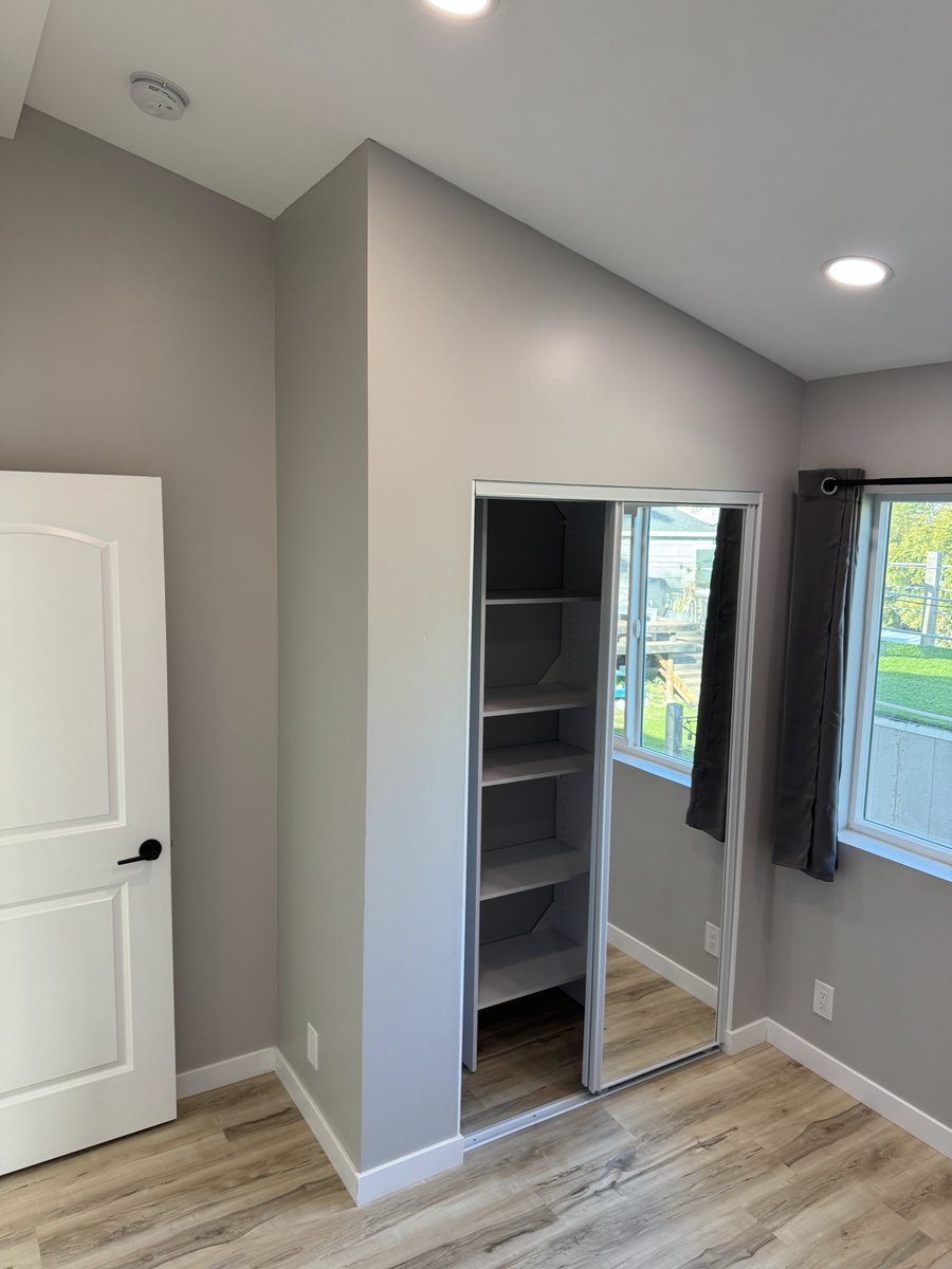 ADU bedroom corner showing a walk-in closet with open shelving, a mirrored sliding door, wood-look flooring, and a window with dark curtains.