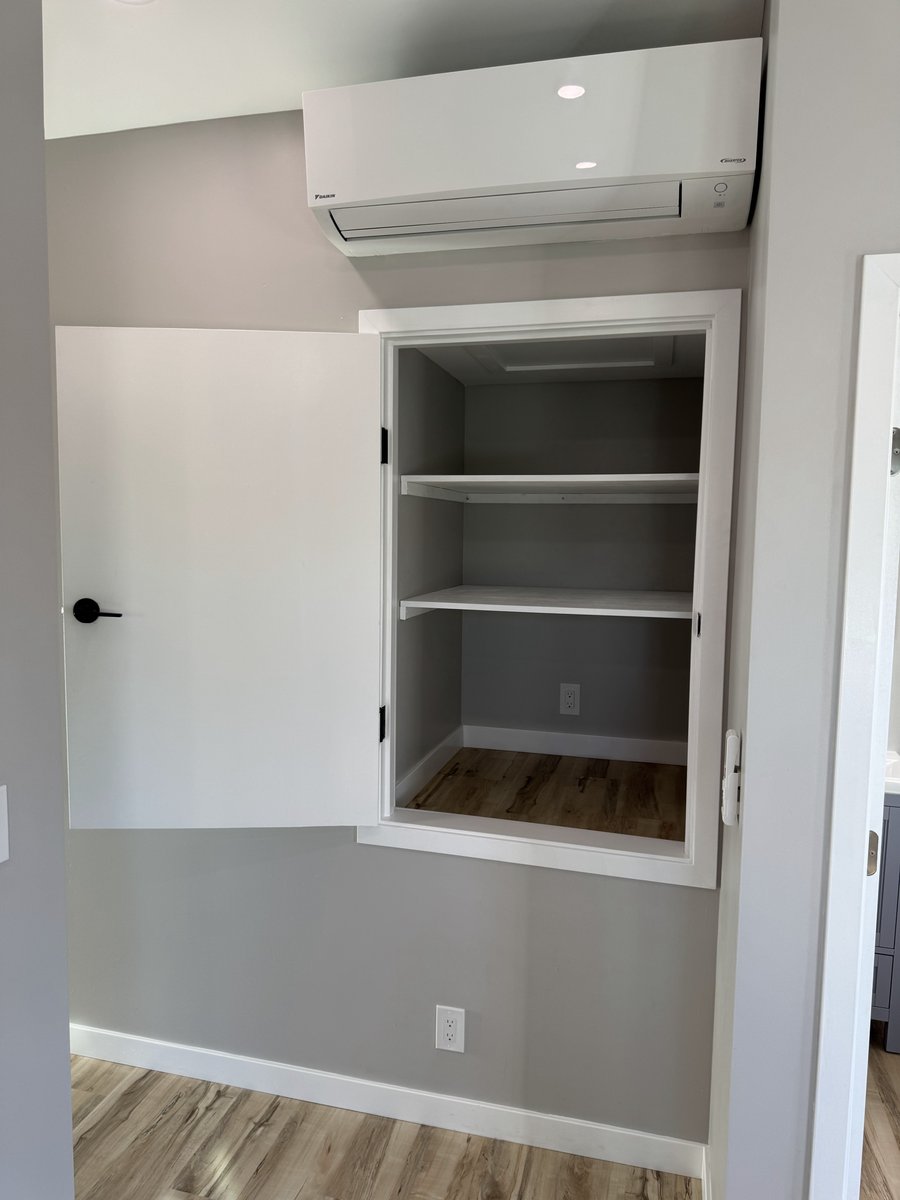 Open built-in wall cabinet with white shelves beneath a mini-split HVAC unit, set against light gray walls and LVP flooring.