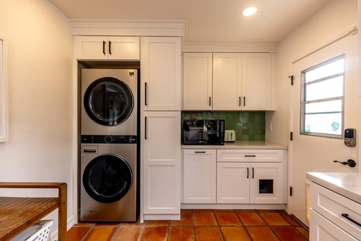 A laundry area within a kitchen, with white cabinets, a stacked washer and dryer set, a green tile backsplash, and terracotta floor tiles.