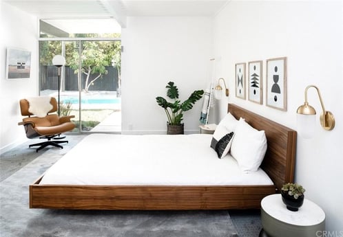 A minimalist bedroom with a large wood-framed bed, white bedding, a tan leather armchair, abstract wall art, and a gold bedside wall sconce.