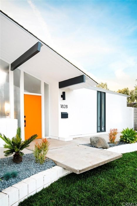 A modern home exterior with a white facade, black trim details, a vibrant orange front door, green landscaping with succulents, and a grass lawn.