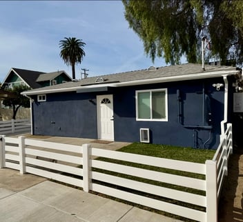 Exterior of a detached ADU with dark blue siding, a white front door, and a white horizontal fence in front, with palm trees in the background.