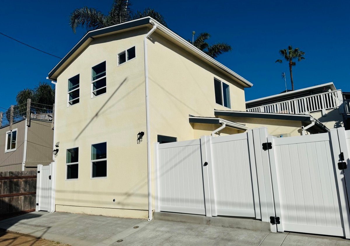 Exterior view of a newly built two-story ADU with light stucco siding, modern black wall sconces, and a white vinyl privacy fence and gate along a concrete driveway, with palm trees in the background.