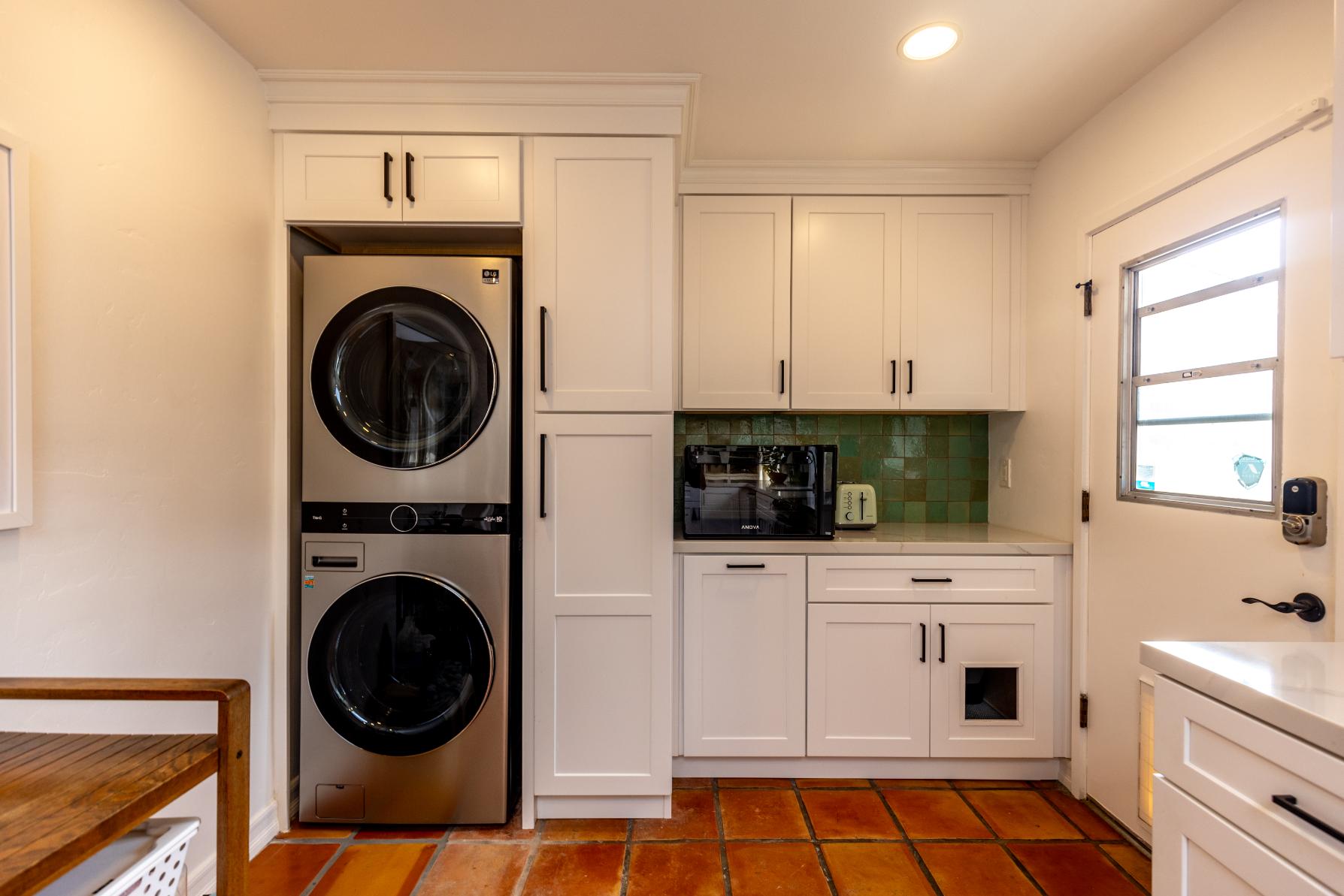 A laundry area within a kitchen, with white cabinets, a stacked washer and dryer set, a green tile backsplash, and terracotta floor tiles.