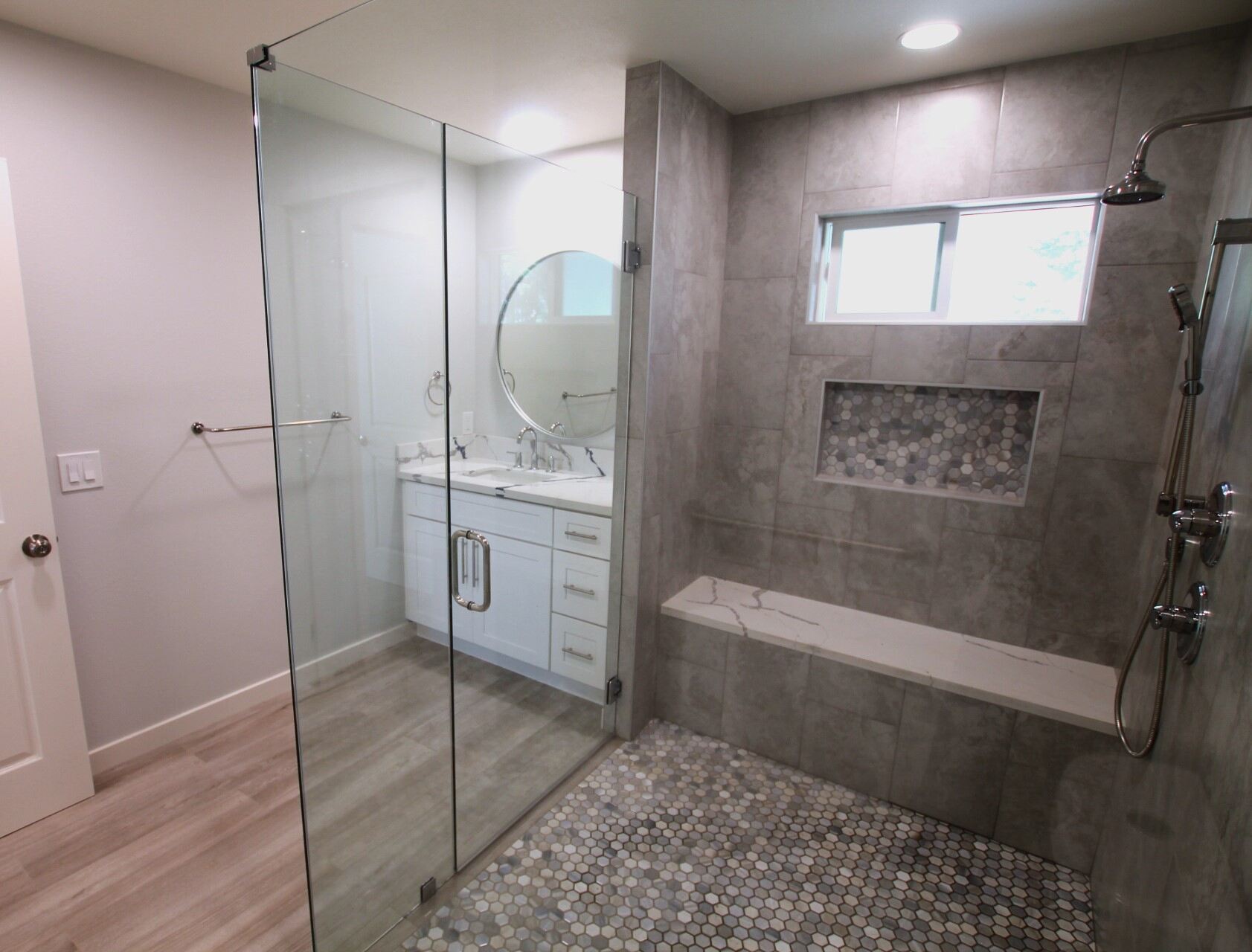 A sleek bathroom with a glass-enclosed walk-in shower, large gray tiles on the walls, a built-in niche with mosaic tiles, and a floating white bench.