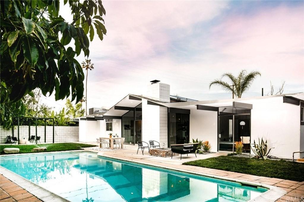 The poolside area includes a small dining set and a couple of lounge chairs on a patio adjacent to a white, single-story house.