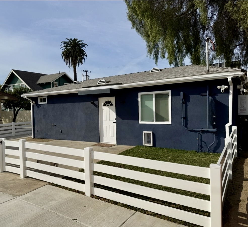 Exterior of a detached ADU with dark blue siding, a white front door, and a white horizontal fence in front, with palm trees in the background.